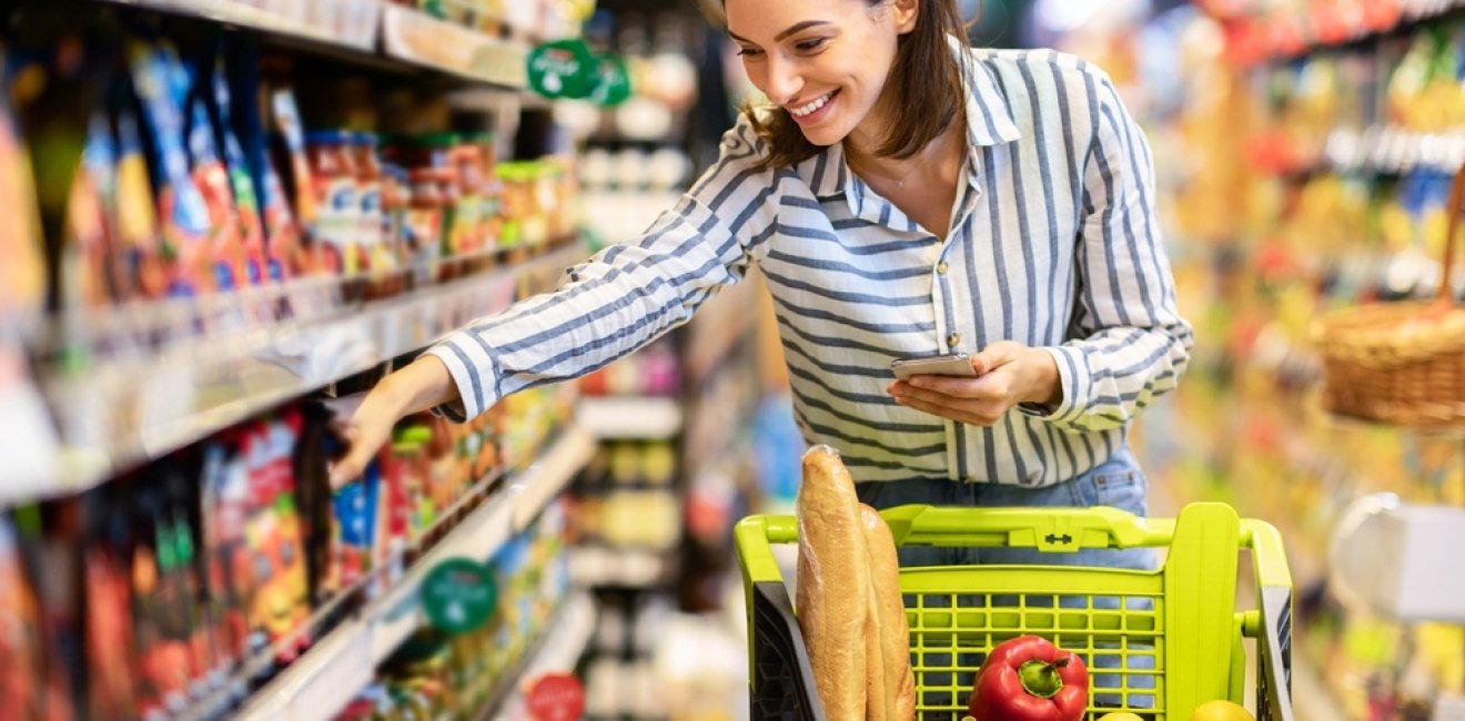 Shopping. Young Smiling Woman Holding And Using Mobile Phone Buying Food Groceries Standing In Supermarket. Female Customer With Smartphone Taking Healthy Products From Shelf At The Shop