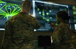 Two soldiers have their back to the camera while looking at a green and blue display of digital information.